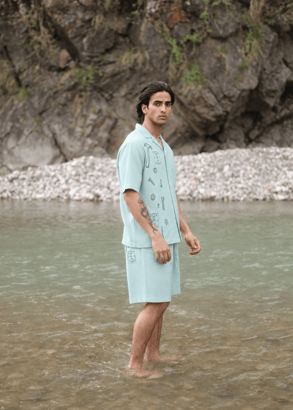 A man stands barefoot in shallow water wearing a light blue short-sleeve button-front camp shirt and matching shorts with dark printed graphics, set against a rocky riverbank background.