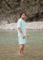 A man stands barefoot in shallow water wearing a light blue short-sleeve button-front camp shirt and matching shorts with dark printed graphics, set against a rocky riverbank background.