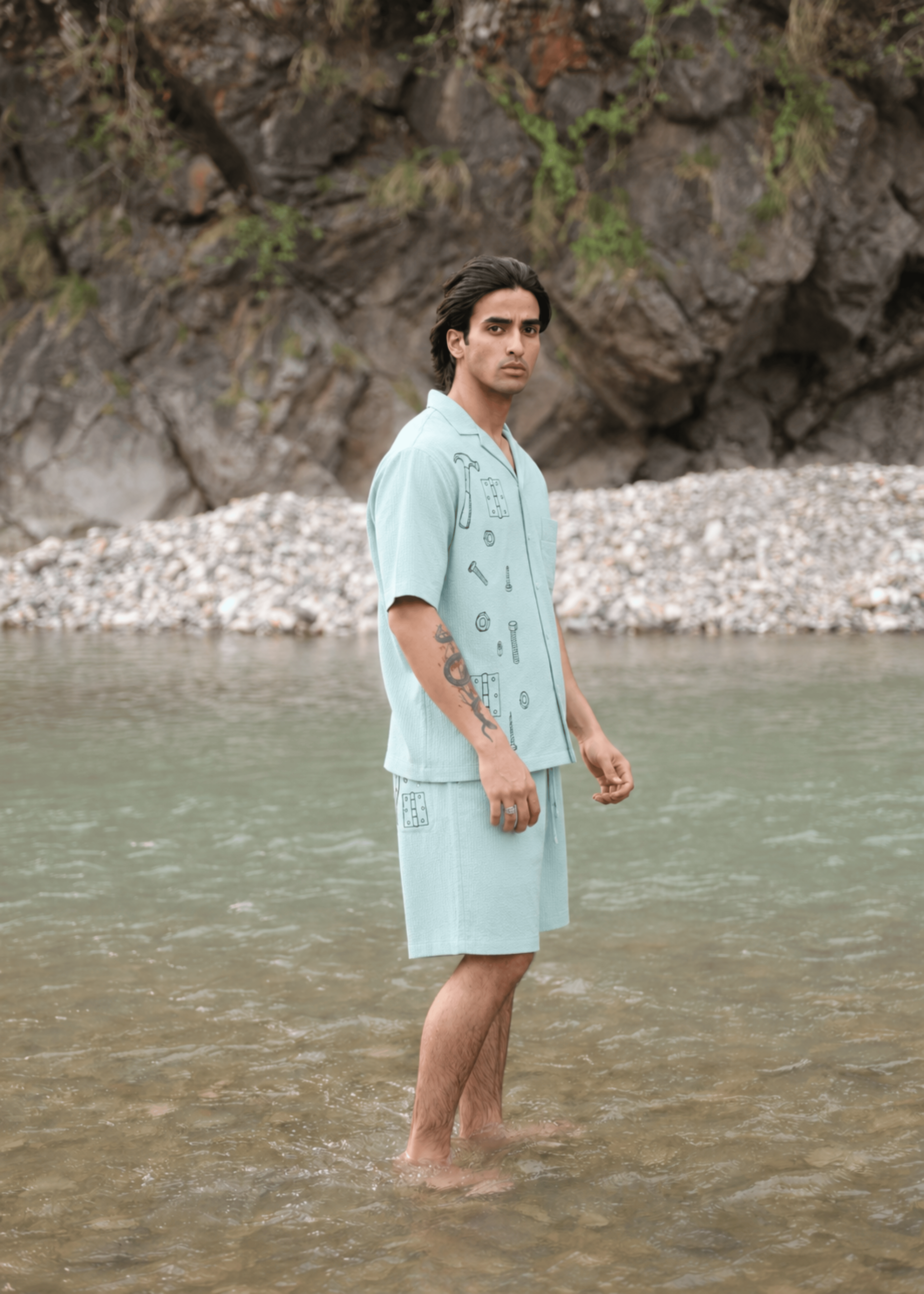 A man stands barefoot in shallow water wearing a light blue short-sleeve button-front camp shirt and matching shorts with dark printed graphics, set against a rocky riverbank background.
